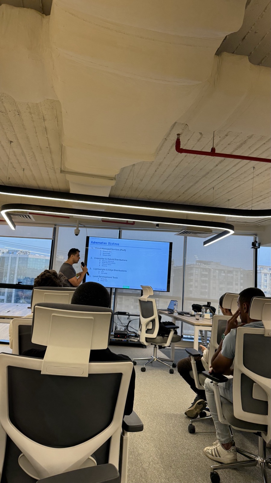 A Kubernetes talk in a glass-walled meeting room, the audience in white chairs